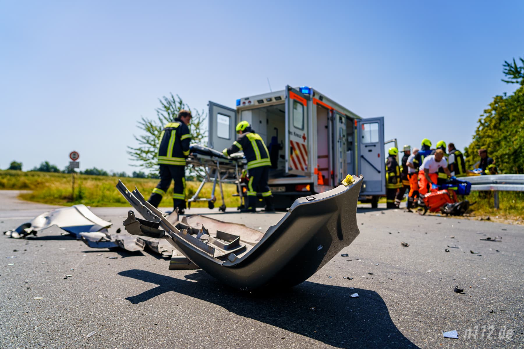 Die abgerissene Frontschürze des Autos liegt auf der Kreuzung - im Hintergrund tragen die Helfer den Verletzten aus dem Gebüsch zum Rettungswagen (Foto: n112.de/Stefan Hillen)